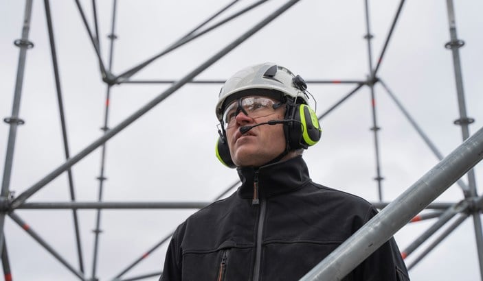 En man i skyddshjälm, hörlurar och headset som tittar ut från ett masttorn.